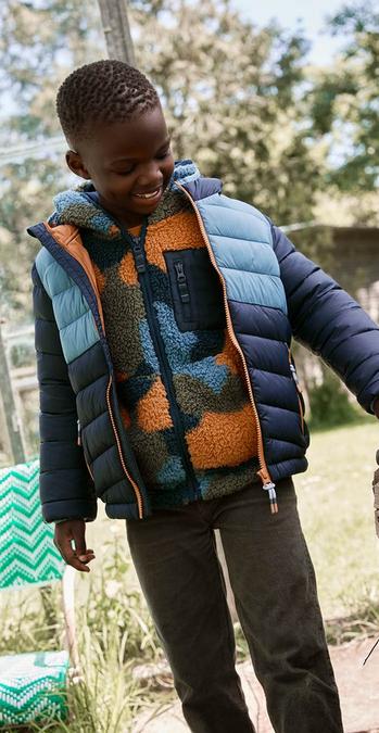 A boy wearing blue contrast colour block padded jacket and orange trim paired with brown trousers.