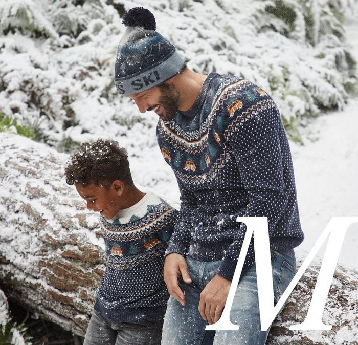 A boy and a man wearing matching campervan patterned Christmas jumpers & jeans, sitting in a snowy forest.