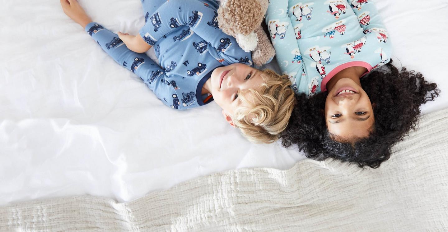 A boy wearing a PJ set with illustrated Land Rover pattern. A girl wearing a PJ set with illustrated penguins pattern.