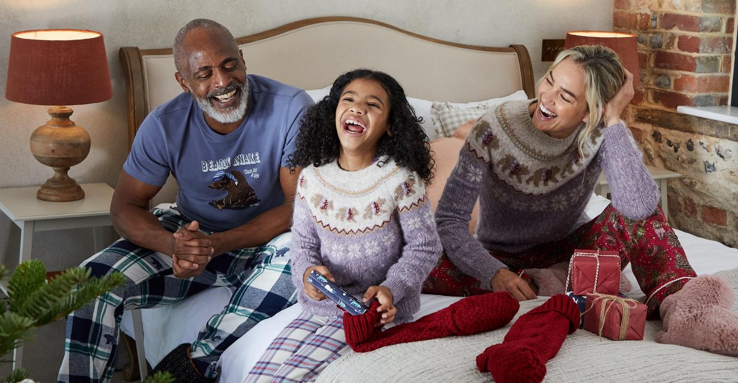 A family dressed in bear character tops & checked PJs, looking at presents together.