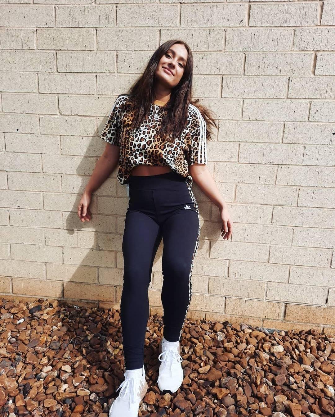 Woman standing against brick wall wearing adidas leopard stipe leggings, a leopard print cropped tee and white adidas NMD running shoes