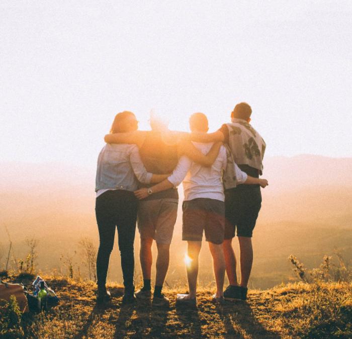 A group of friends with their arms around each other, at the top of a hill at sunrise looking down at the views
