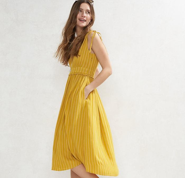 A brown-haired model wearing a FatFace stripe midi dress in yellow with black sandals.