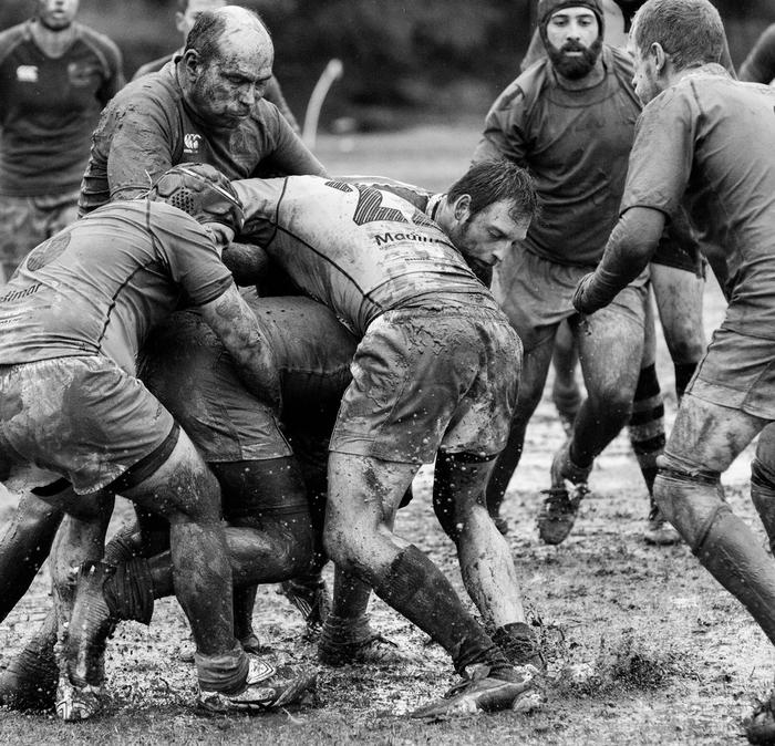 A group of muddy rugby players on a water-logged field mid-scrum