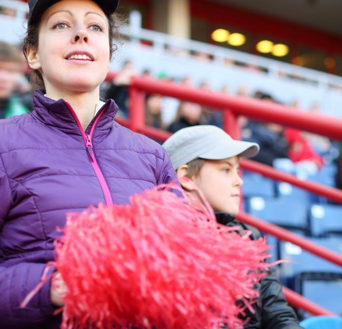 A woman and a young boy looking on from a football stand, holding a red pom pom. 