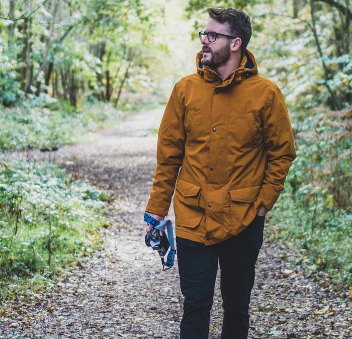 Man walking through a forest in a yellow coat and wearing glasses holding a camera