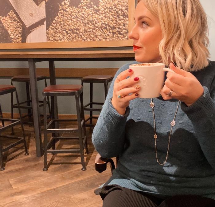 Blonde women holding a cup of hot chocolate sat in a café wearing a teal knitted jumper and long necklace