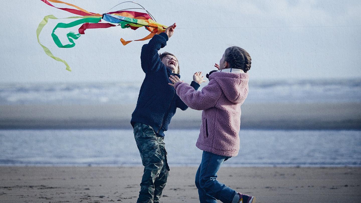 Two children playing on a beach with a kite. One child is wearing a pink coat with blue jeans and the other is wearing a navy coat with camo trousers.