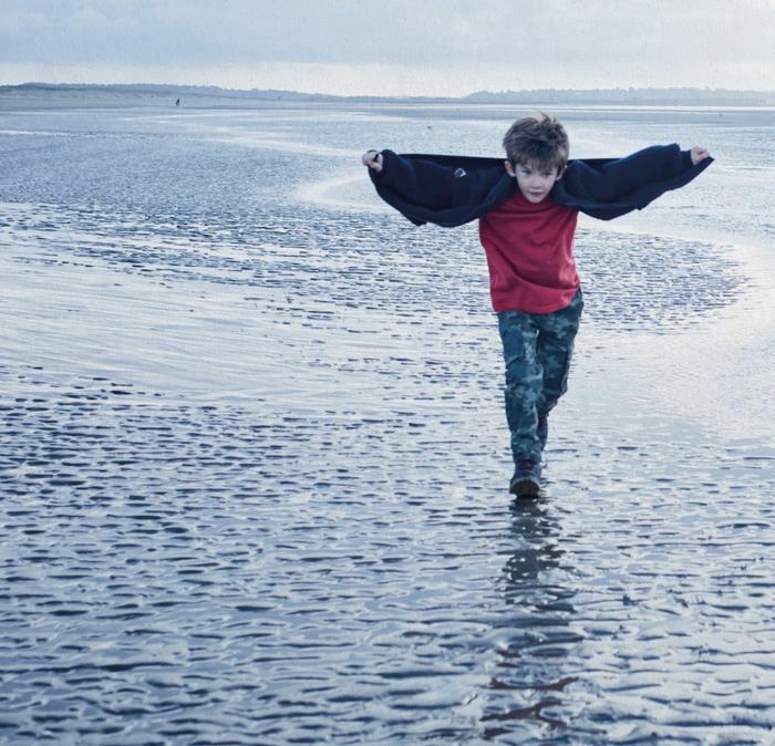 A young boy playfully running along the beach, his jacket open like a cape.