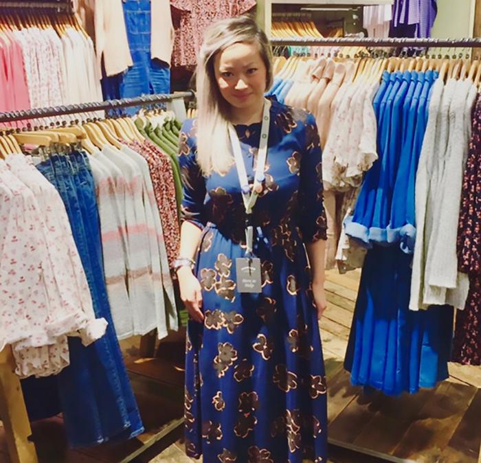 A woman stood in front of a rail of clothes in a shop wearing a navy floral wrap dress with a staff lanyard around her neck.