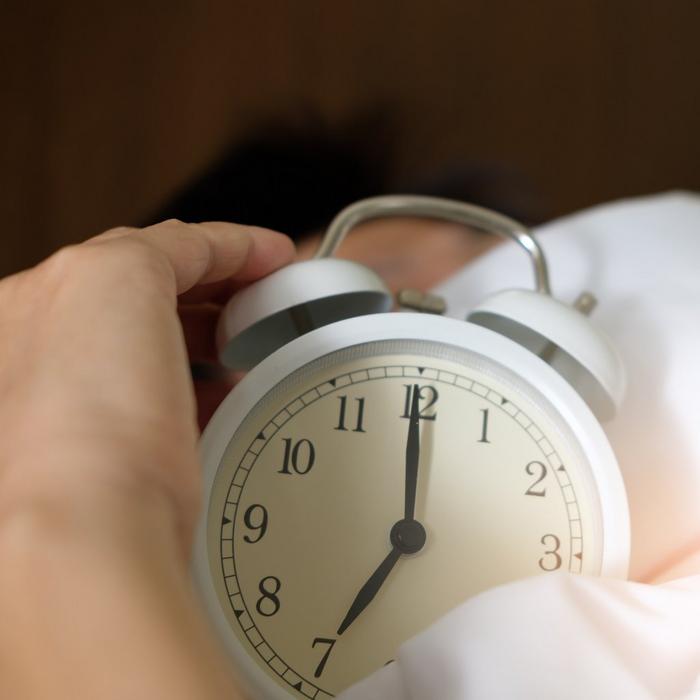 A person in bed snoozing an alarm clock.