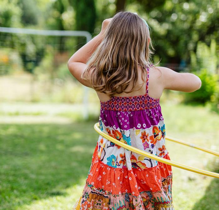 A girl wearing a bright summery dress playing with a yellow hula hoop in a green garden.