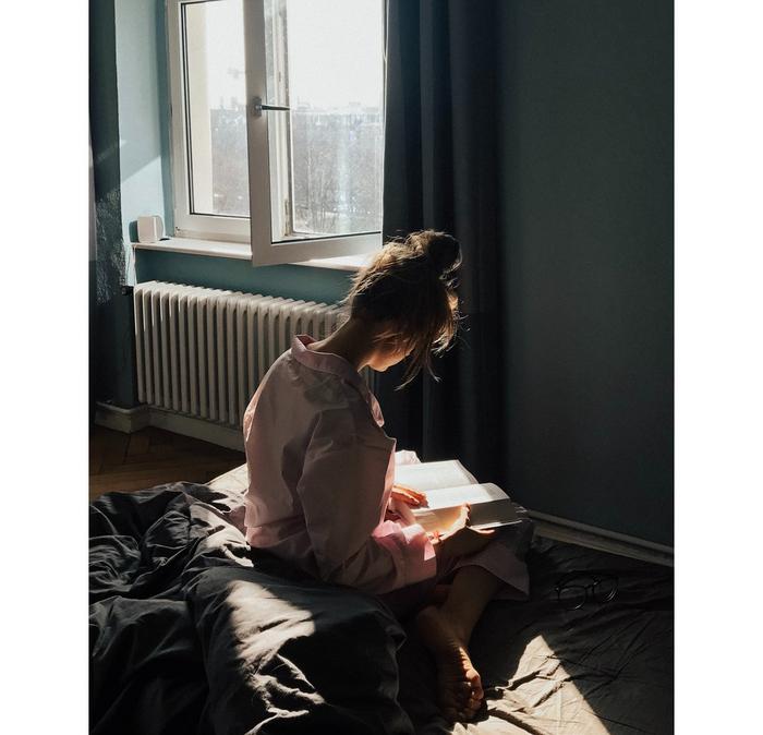 A woman sitting on a bed, reading a book in the natural light from the window.