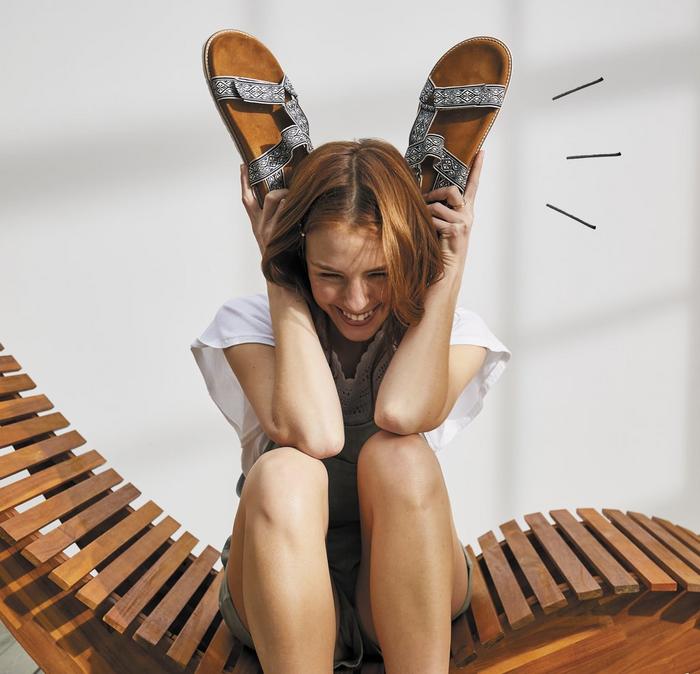 A woman sitting on a wooden sun lounger holding a pair of sandals up like bunny ears.