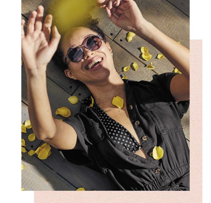 A woman wearing sunglasses & a black playsuit surrounded by yellow petals.