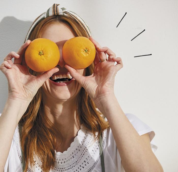 A woman making a silly face by holding a pair of oranges over her eyes.