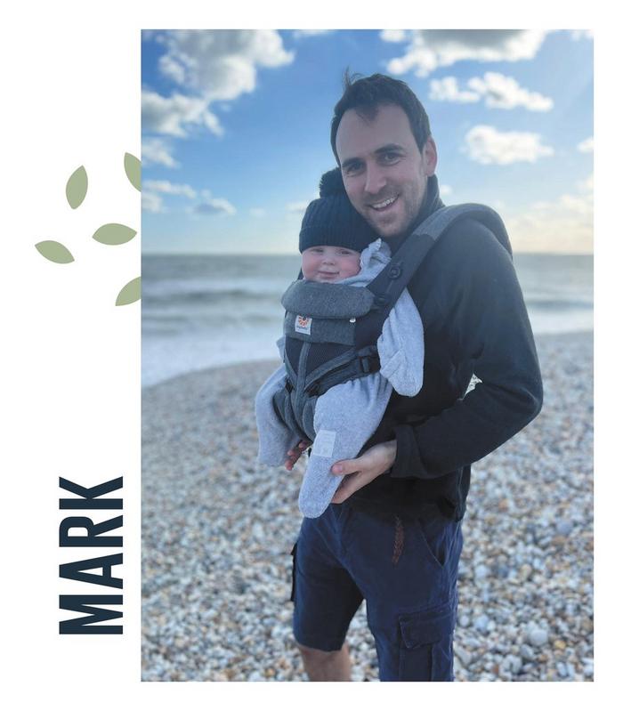 A family photo featuring dad Mark & his baby boy, smiling on a pebbled beach.