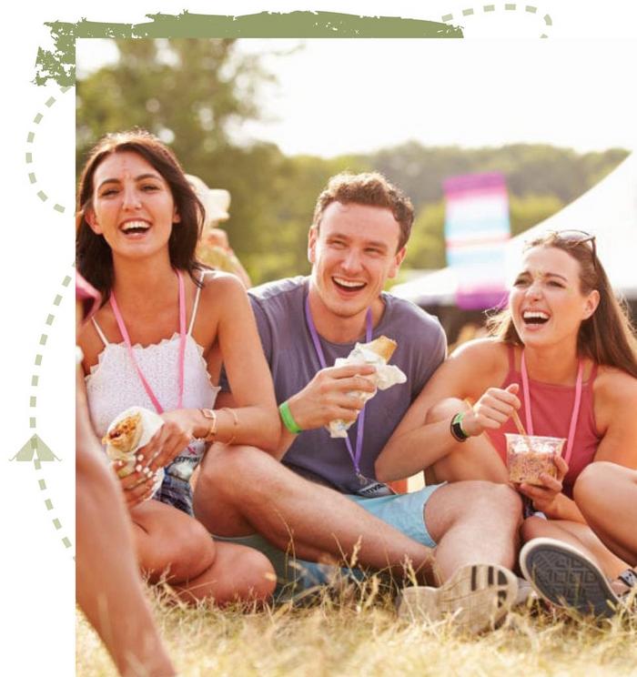 A group of friends wearing summer tops & shorts, eating together in a field at a festival.