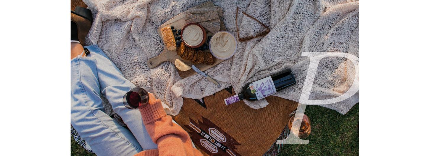A woman wearing a pink jumper & blue jeans, holding a glass of red wine & enjoying a picnic on a blanket.
