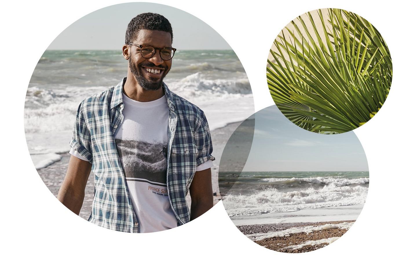 A man in a check shirt & graphic t-shirt, smiling by the sea. Spiky palm leaves. Waves crashing on a beach.