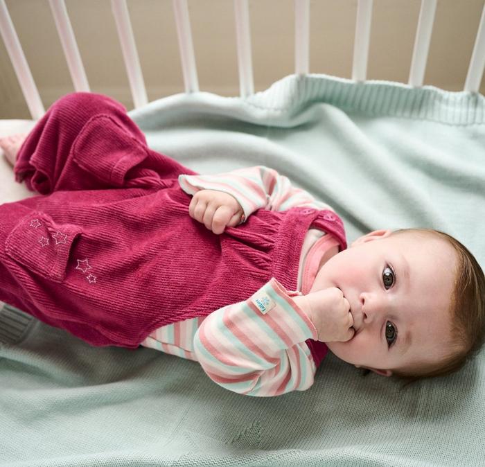 A baby girl in a crib, wearing dark pink soft cord dungarees over a long-sleeved pastel striped T-shirt.