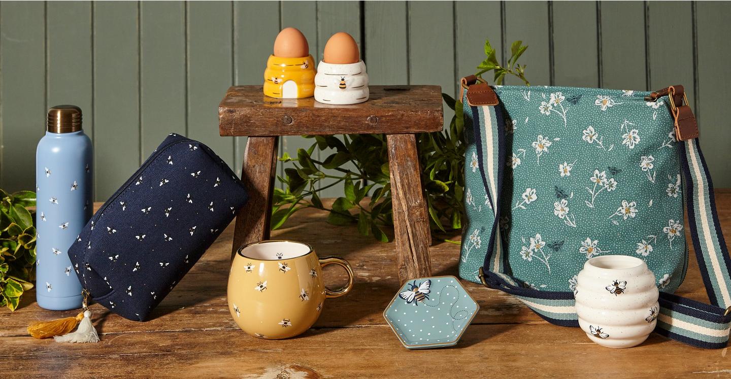 A collection of bee-themed products, including a water bottle, wash bag, ceramic mug, hive-shaped egg cups & candle, & a bag.