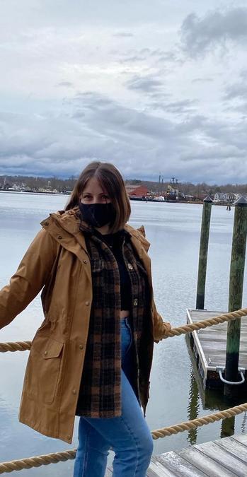 A woman standing on a jetty by a harbour, wearing a mustard yellow waterproof jacket, check scarf, black top & blue jeans.