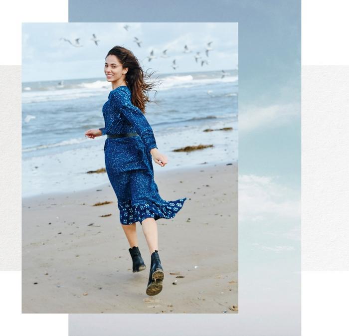 A woman running across a beach towards the sea, wearing a tiered blue dress with spot & floral prints, & black ankle boots.