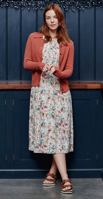 Woman wearing a wide-collared salmon-pink knitted cardigan over a shirred white dress with subtly multi-coloured floral print.