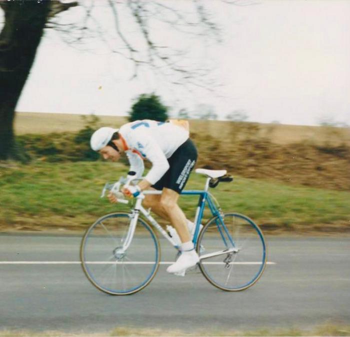 A man in full cycling gear & helmet riding a classic Raleigh road bike along a country road.