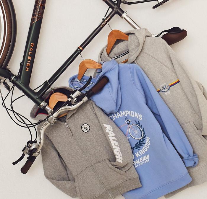 3 hoodies in pale grey & powder blue, each with Raleigh graphics, hang from the frame of a Raleigh bicycle.