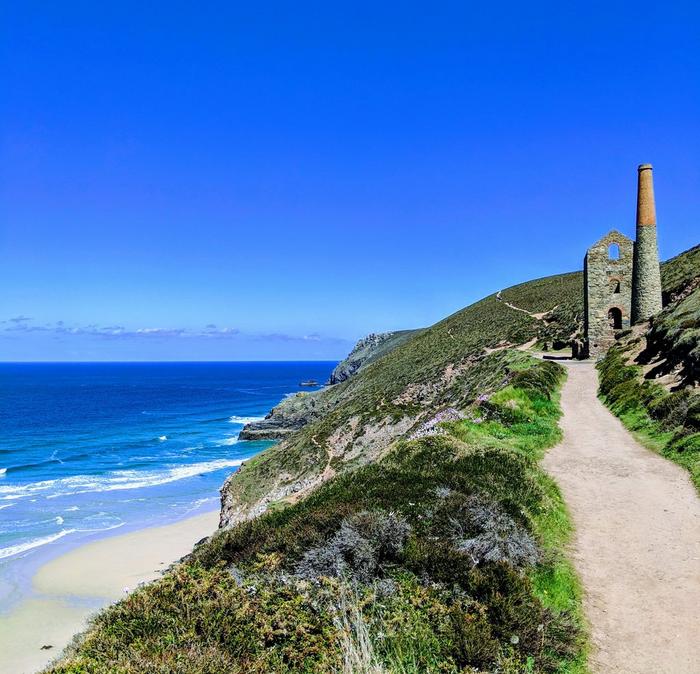 A coastal foot path leads to the remains of an old stone building & chimney overlooking the sea.