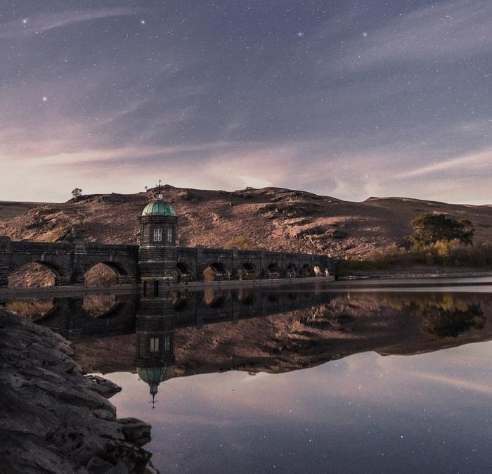 A historic stone bridge frames the edge of a perfectly still reservoir, reflecting the sky full of stars above.