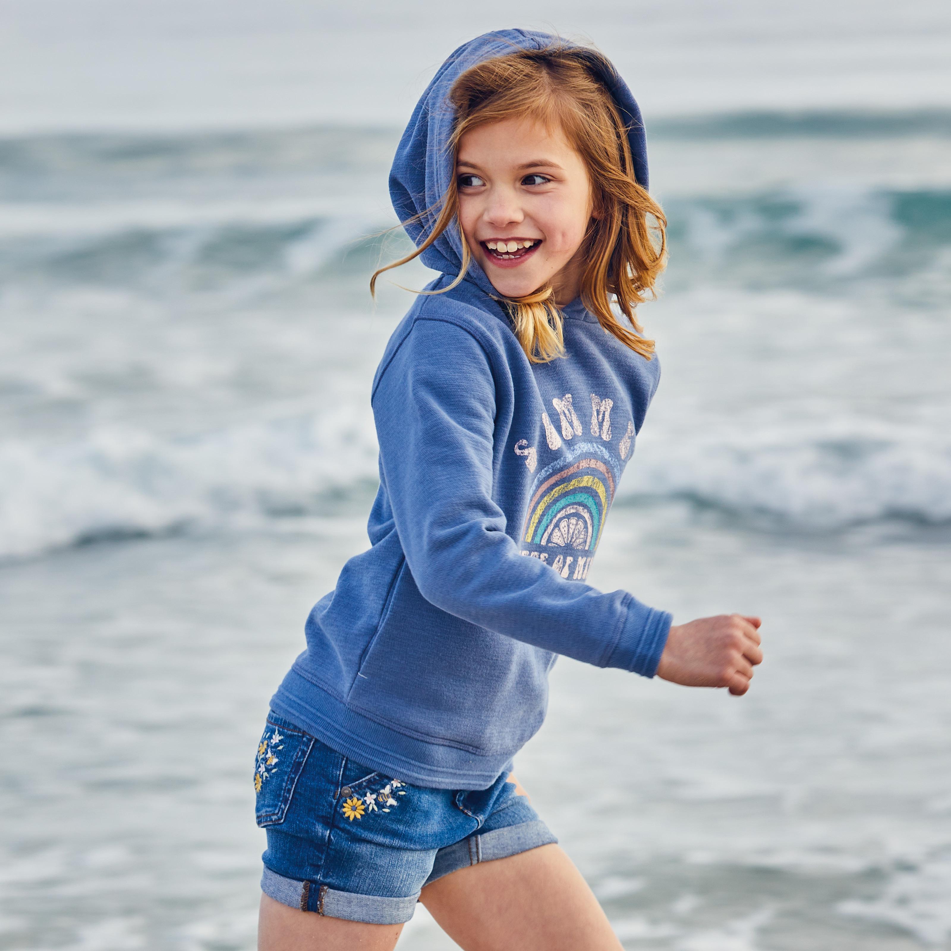 Girl wearing a blue popover hoodie with retro-style Summer graphic on the front, & denim shorts with embroidered flowers.
