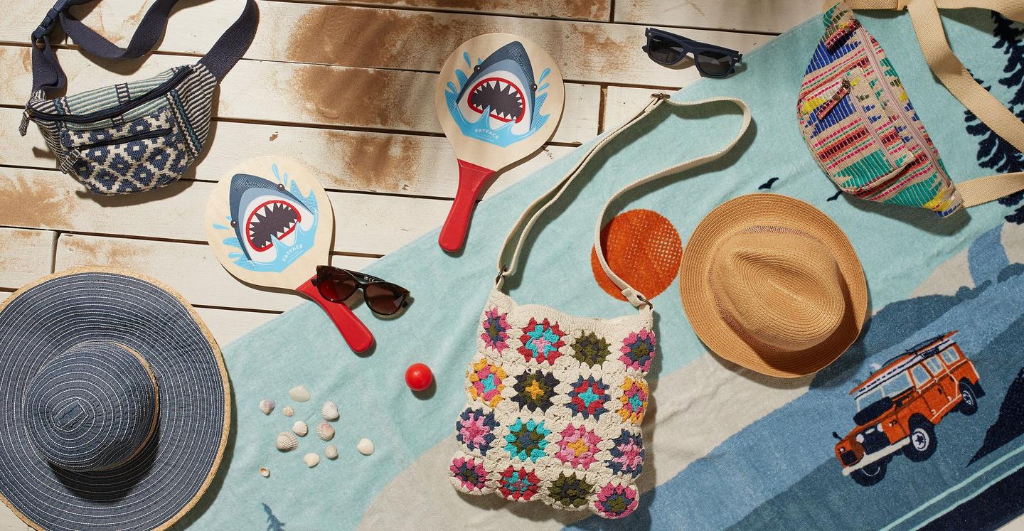 A range of accessories on a beach towel, including geo patterned bumbags, straw sun hats, sunglasses, bag, tennis bats & ball.