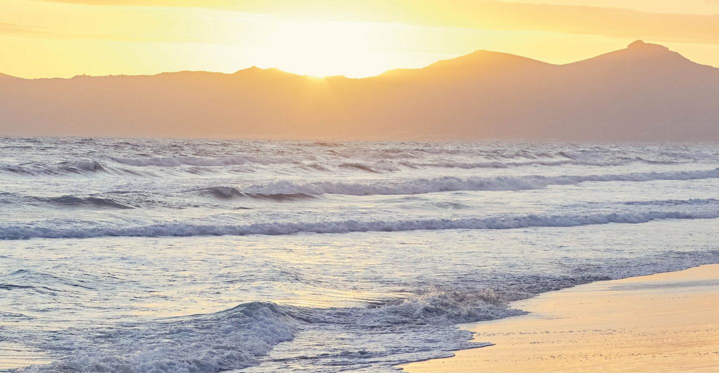 Waves gently lapping at a sandy beach, as the sun sets over distant hills.