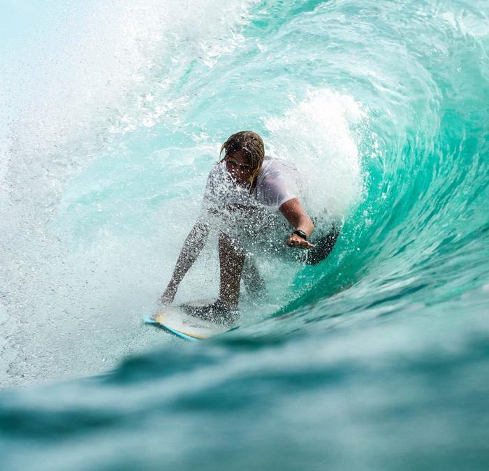 A surfer in soaked T-shirt & shorts, riding in the tube of a crystal clear turquoise-green wave.