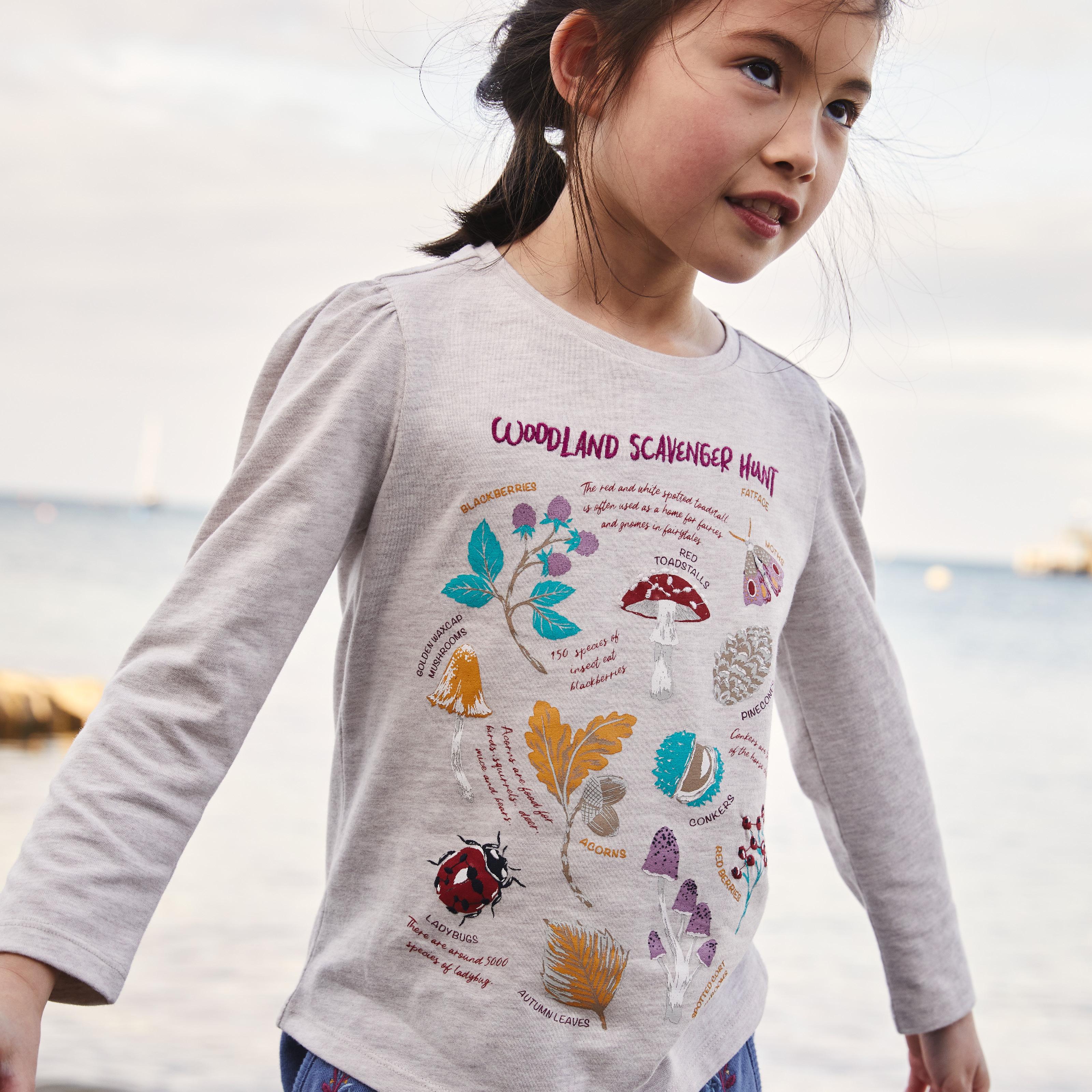 Girl wearing a long-sleeved ecru-coloured T-shirt with 'Woodland Scavenger Hunt' illustrations of insects & plants.
