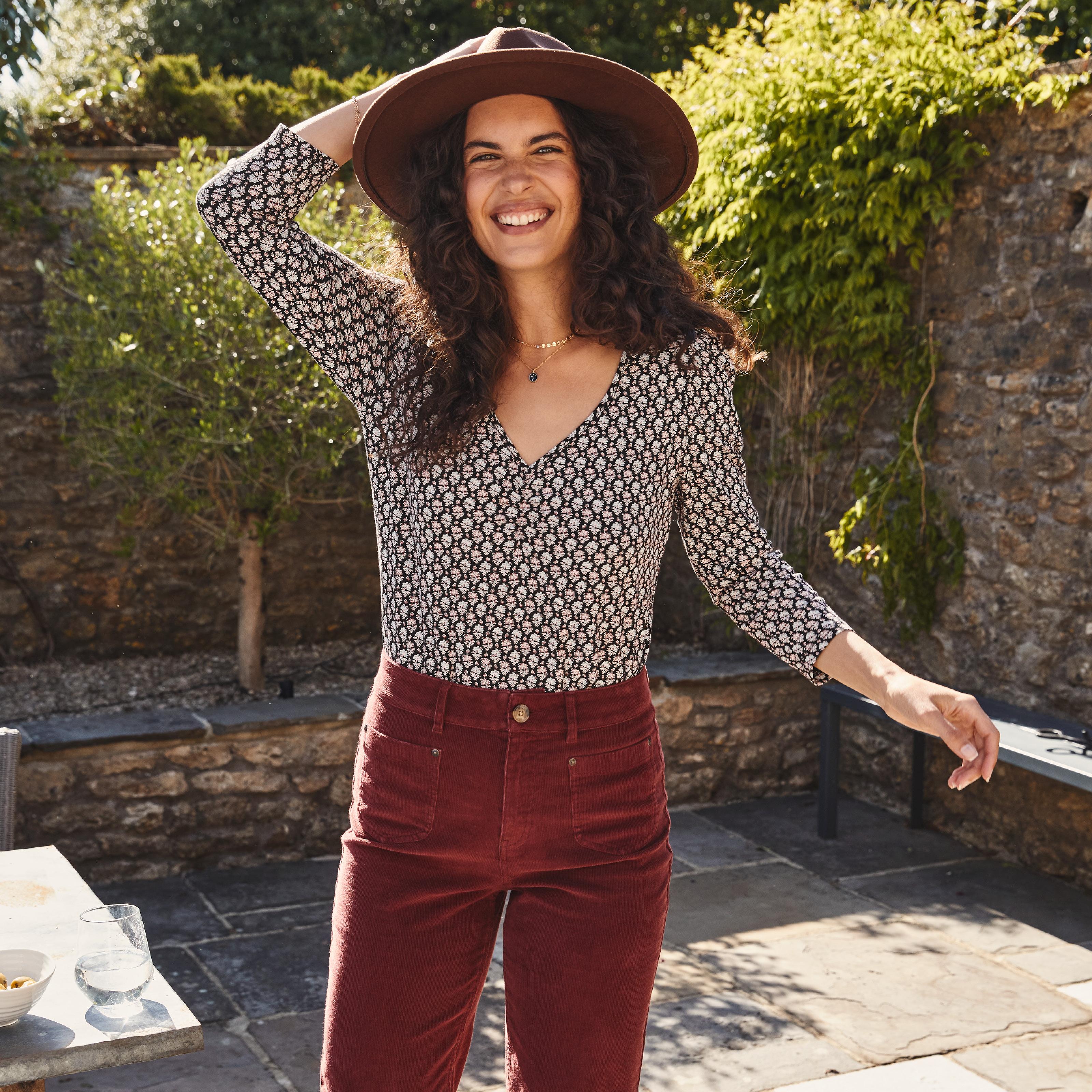 Woman wearing a wide brimmed brown hat, floral tile print top with 3/4 length sleeves, & red cord trousers.