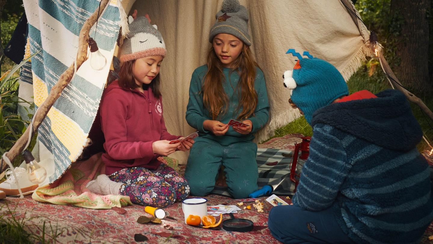 3 children wearing warm hats & cozy clothes, playing a card game in an outdoor blanket fort.