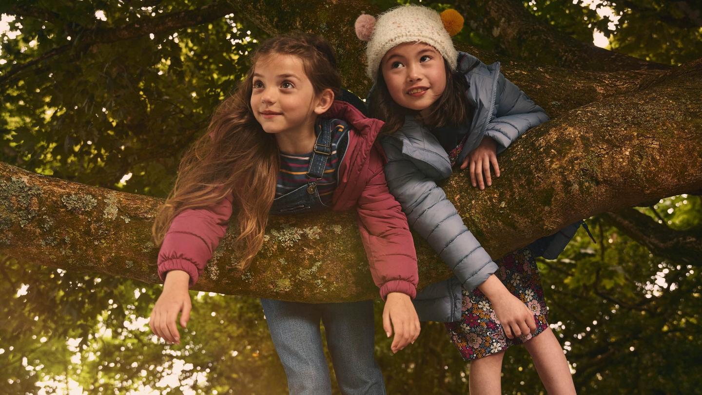 Two girls wearing puffer jackets & colorful outfits, up in a tree.