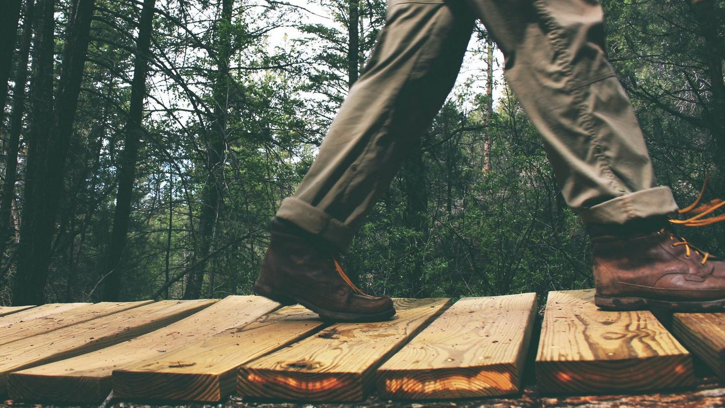 A person wearing khaki green utility trousers & brown leather boots, crossing an uneven walkway in a forest.