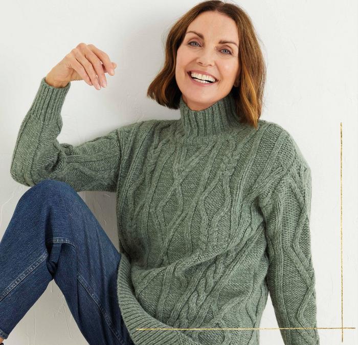 A woman wearing a green chunky cable knit jumper with high neck, & blue jeans.
