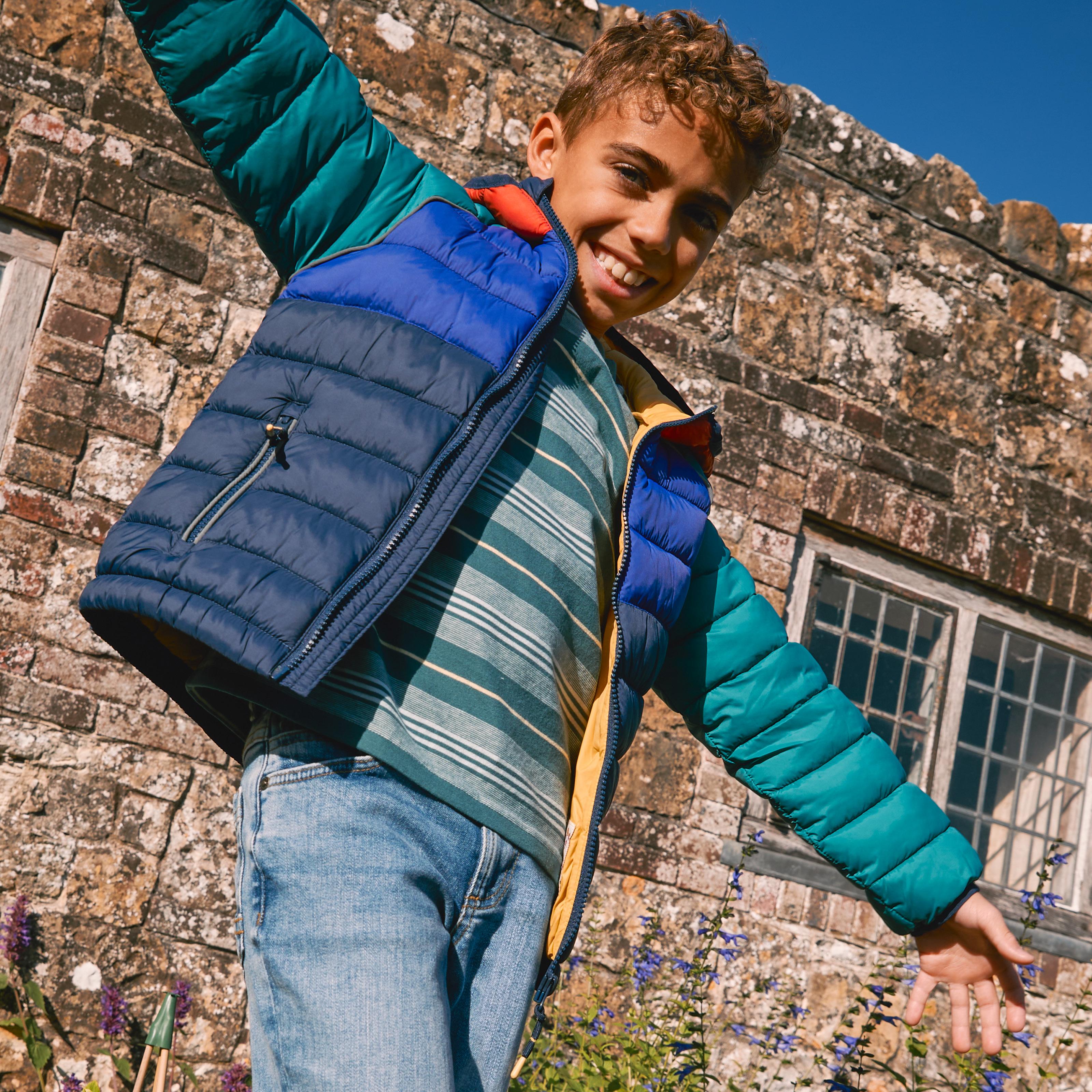 A boy wearing a multi-coloured colour block puffer jacket, over a teal & white striped T-shirt & light blue jeans.