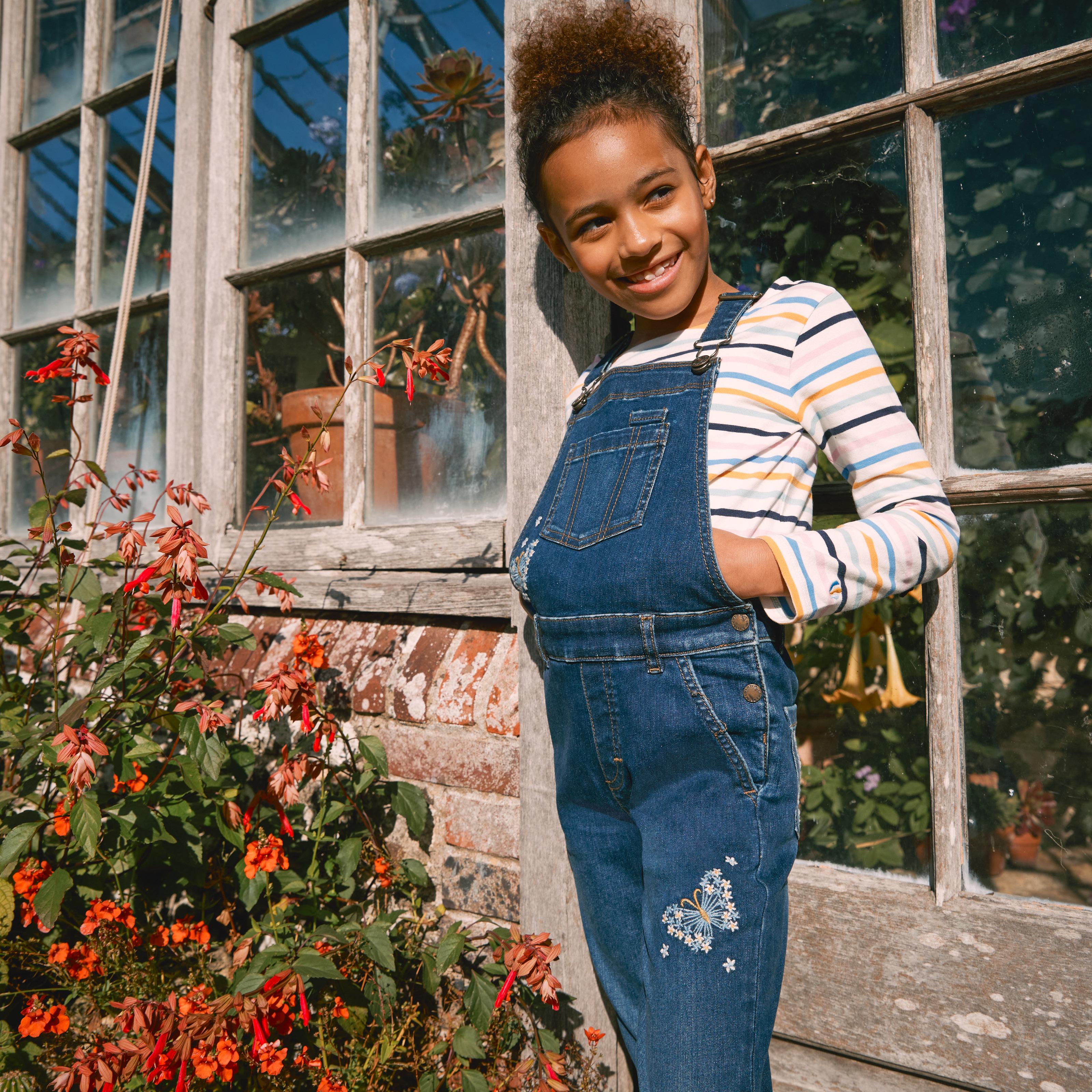 Girl wearing blue denim dungarees with floral butterfly embroidery, over a white T-shirt with colourful Breton-style stripes.