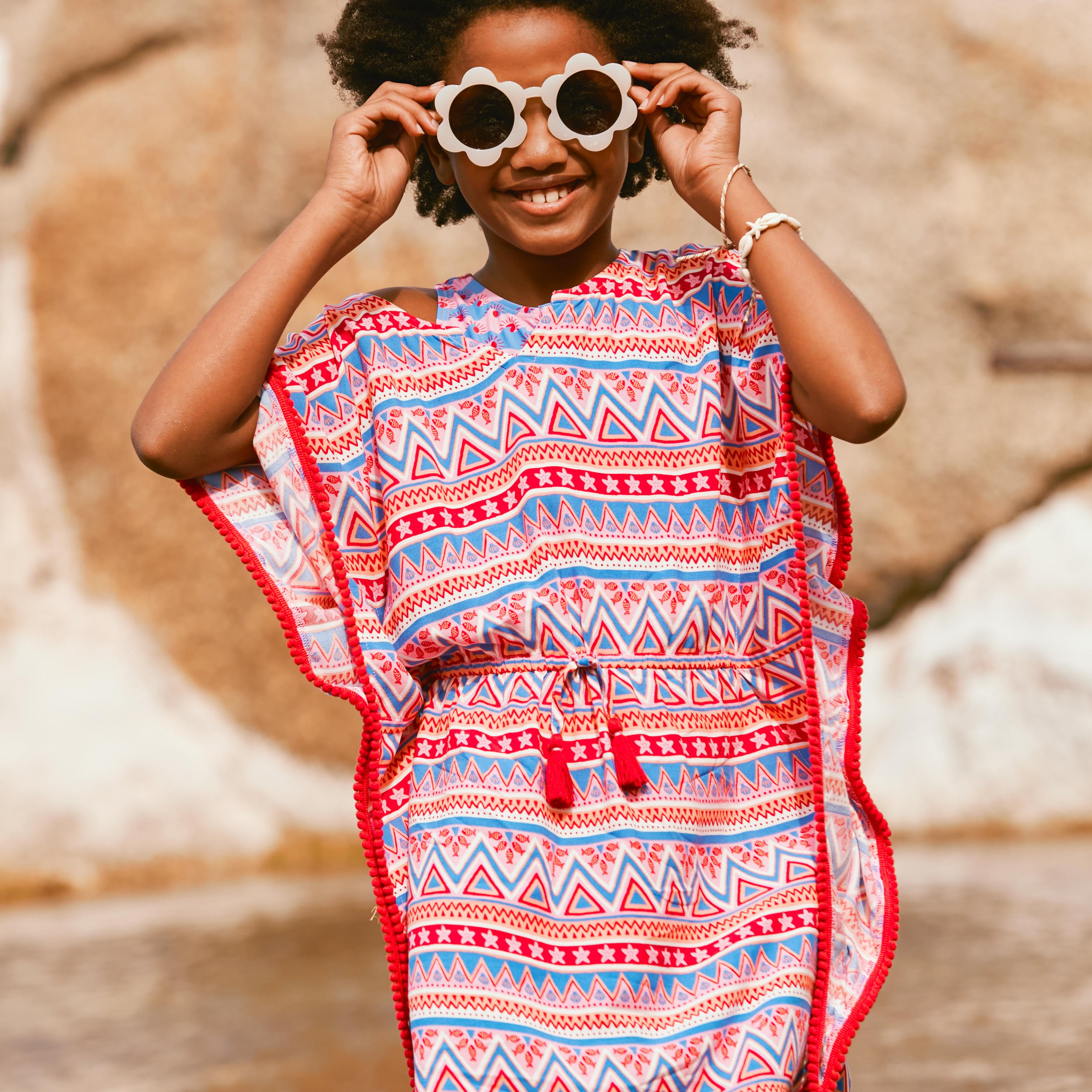 A girl wearing flower-shaped sunglasses & a brightly coloured kaftan with blue & red geometric prints & tie waist.