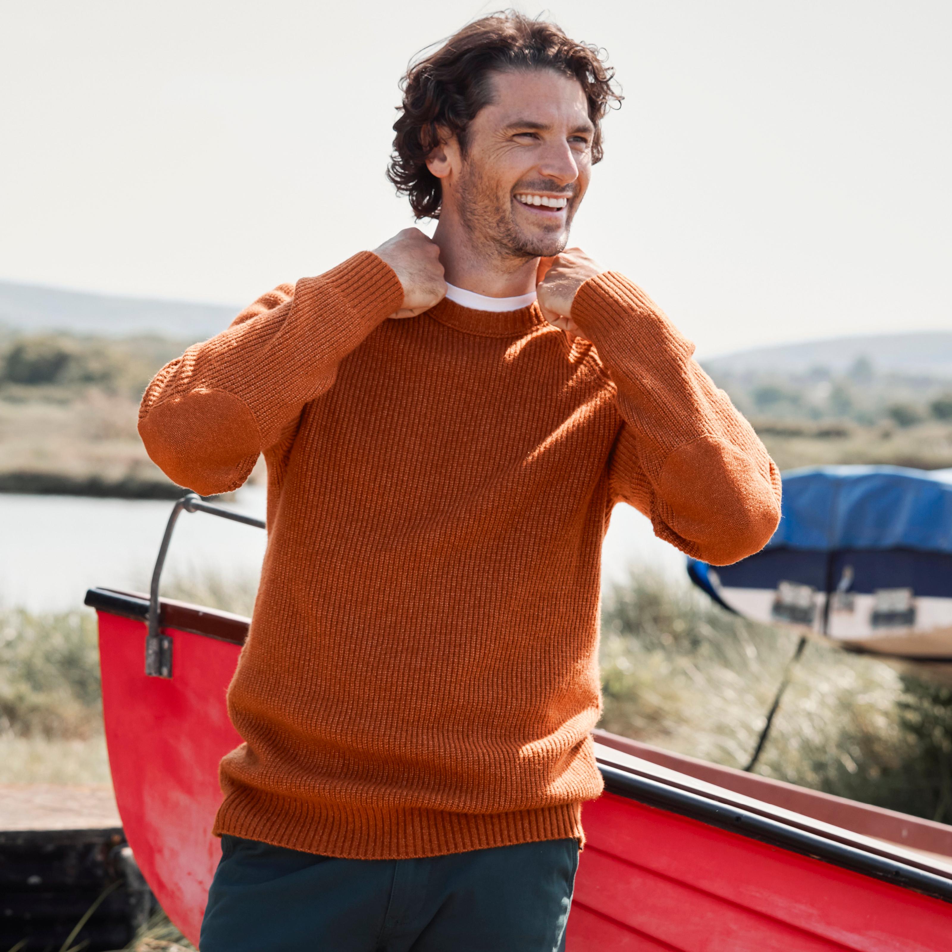 A man wearing a dark orange ribbed-knit jumper over a white T-shirt & dark blue trousers.