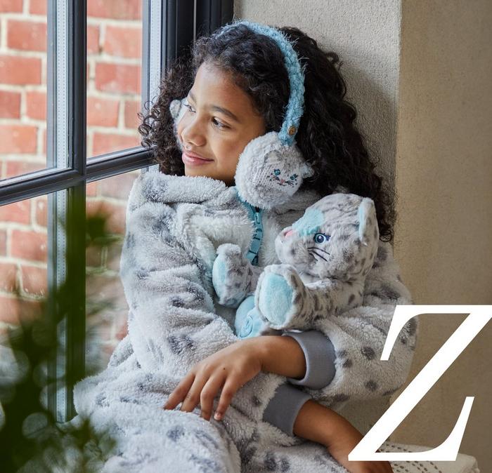 A girl wearing a Sadie the Snow Leopard character onesie with matching ear muffs & Sadie plush toy.