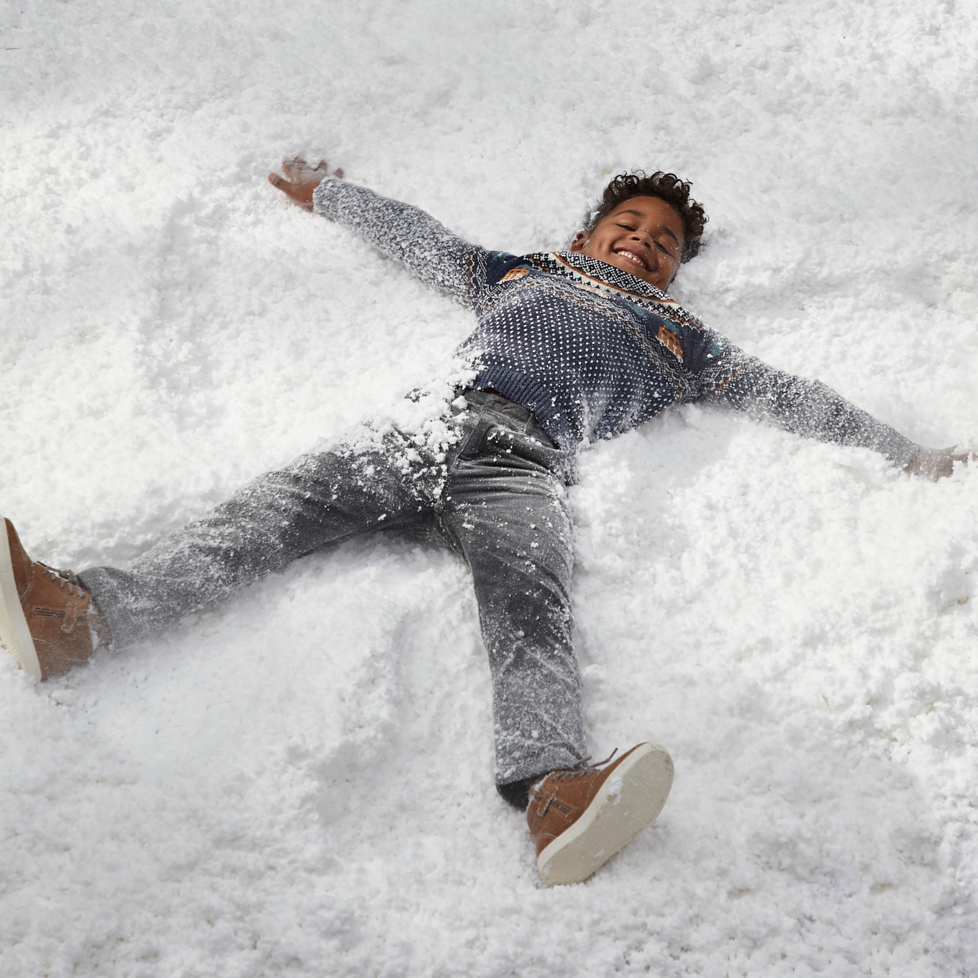 A boy wearing a navy blue jumper with festive campervan pattern, & grey jeans, making a snow angel.