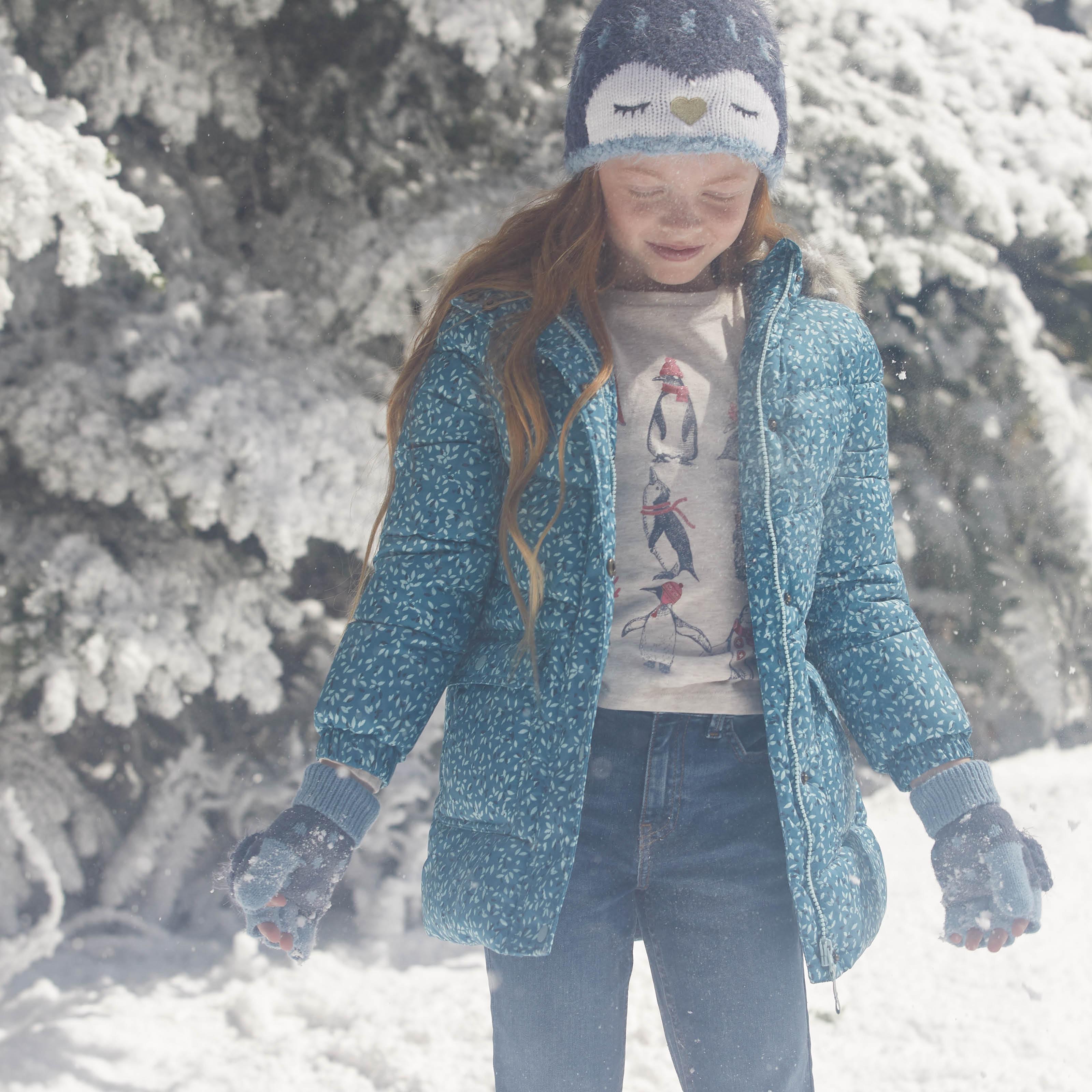 A girl wearing a penguin character beanie hat & matching gloves, & a bright blue patterned coat over a T-shirt & blue jeans.